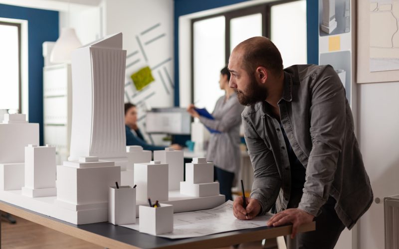 Man working on architectural design at office while looking at laptop computer for industry construction. Project manager inspecting maquette modern design building model